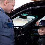 a young girl wears the hat of a state patrol officer sitting in the driver seat of the patrol car while sargent smiles off to the left side